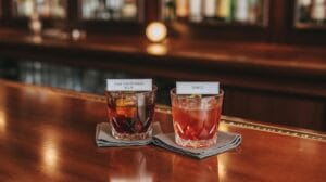 Two cocktails in short glasses sit on napkins at a bar. The drink on the left is labeled Old Fashioned Sour and the one on the right is labeled Spritz. The background shows a blurred bar setting.