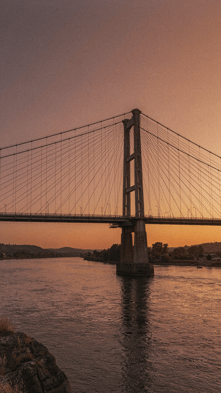 A large suspension bridge stretches across a river at sunset.