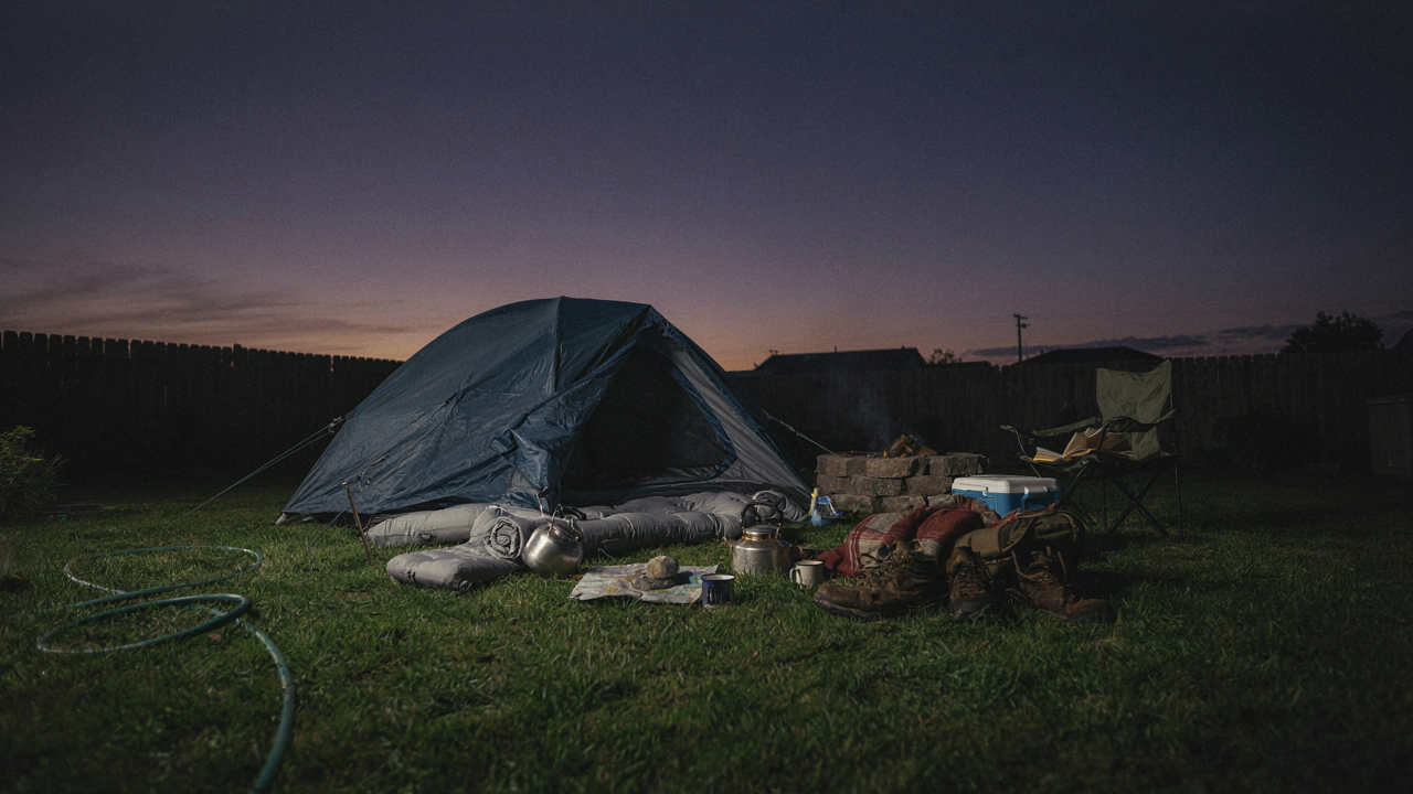 A messy backyard campout setup at dusk with a tent and gear.