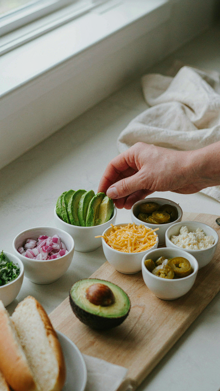 A hand reaches for a bowl of fresh avocado slices, a hot dog topping.