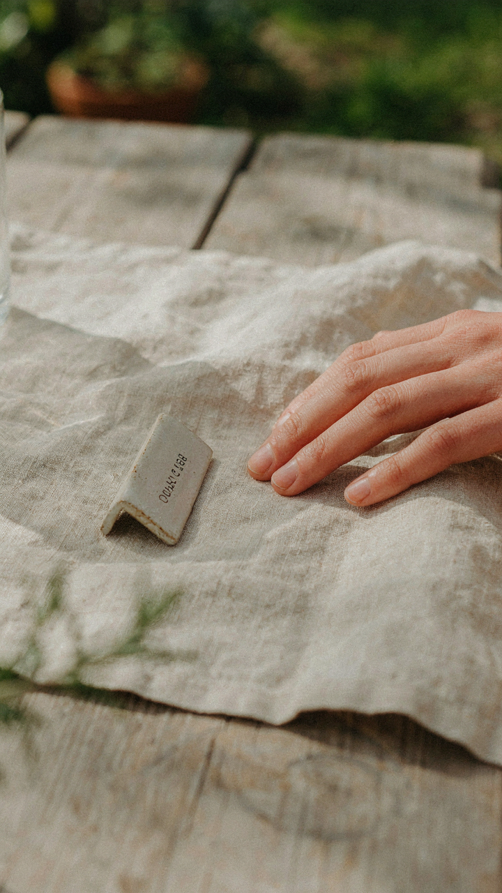 A hand touches a rough linen napkin next to a ceramic outdoor dinner place card.