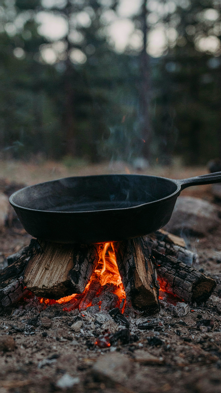 A large cast iron skillet sits over glowing campfire embers.