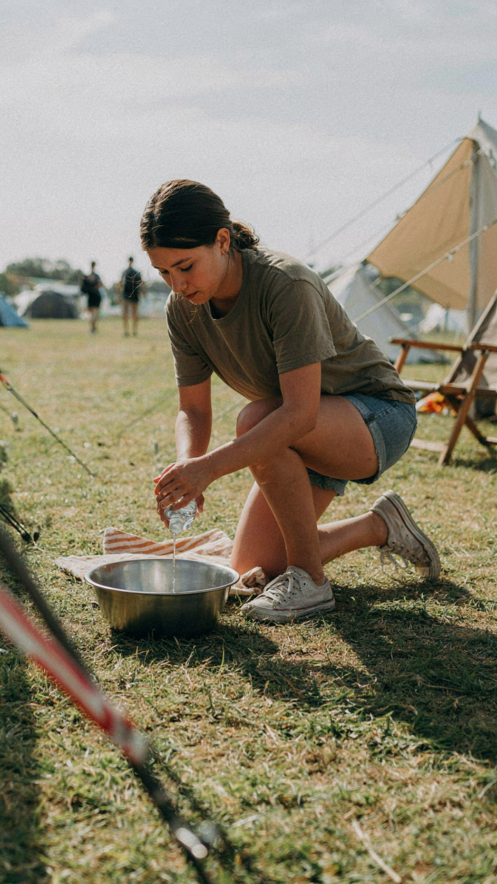 A person pours water from a bottle into a small basin at a festival campsite.