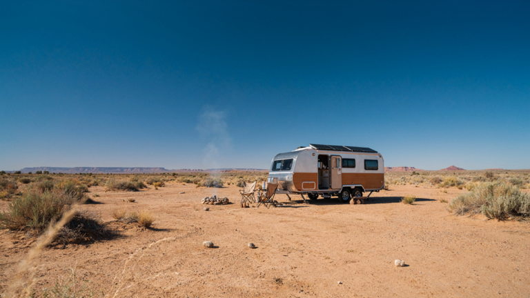 A white camper trailer with solar panels on its roof sits in a sunny desert.