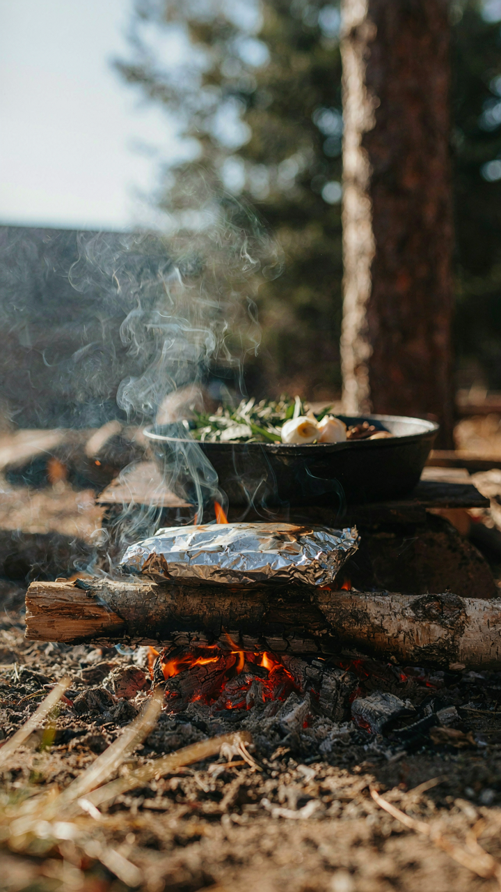 Smoke curls around a foil packet cooking on campfire embers.