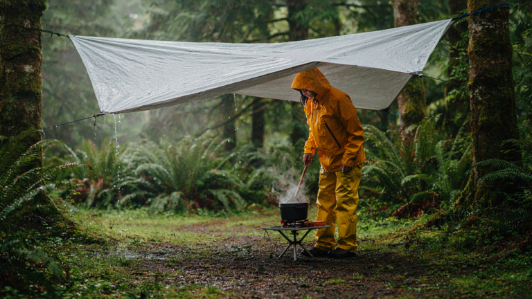 A person cooks under a tarp in a rainy forest, demonstrating camping rainy day hacks.