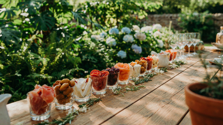 An array of charcuterie cups sit on a long wooden table at an outdoor garden party.