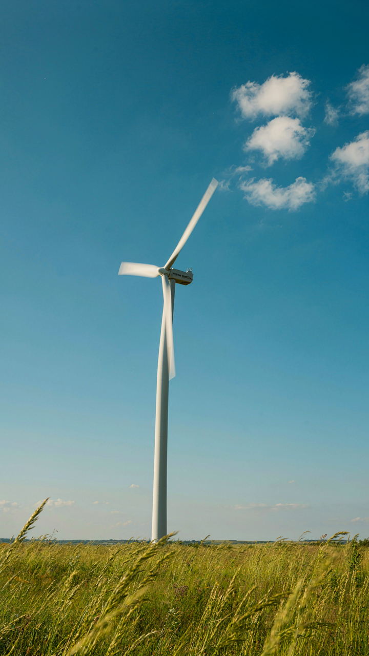A wind turbine spins fast against a blue sky with tall grass blowing.