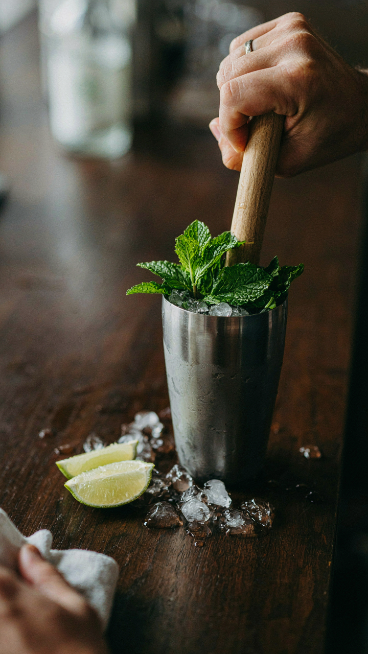 A bartender's hand muddles fresh mint leaves in a cocktail shaker on a bar.