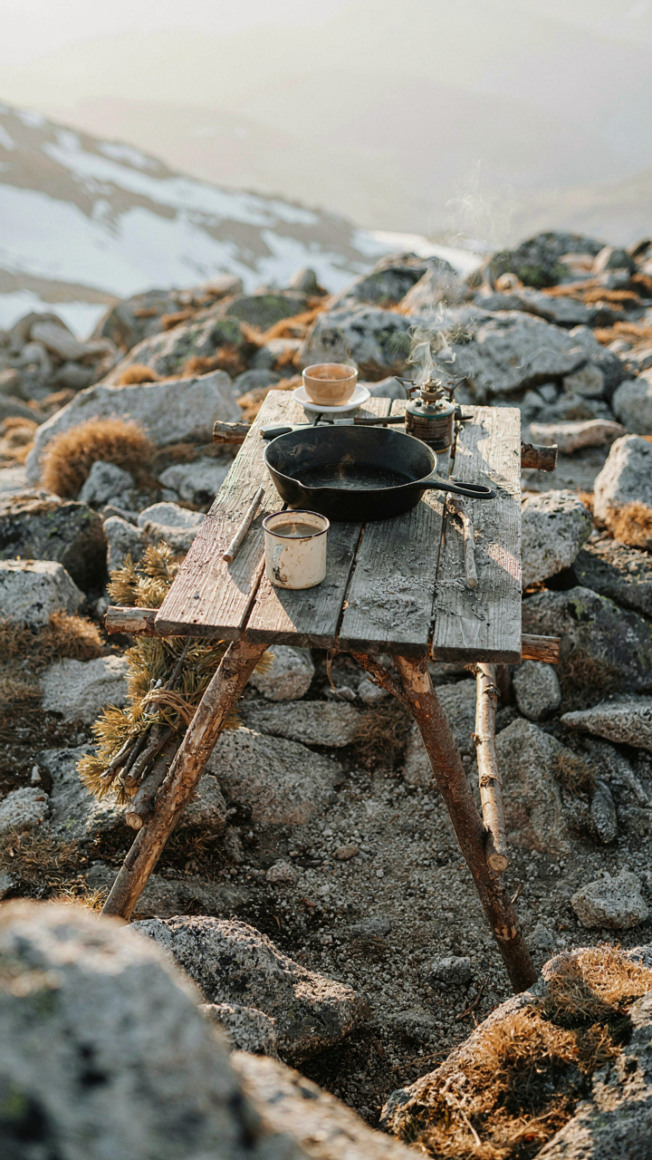 A sturdy diy camp table holds a heavy cast iron skillet on rocky, uneven ground.
