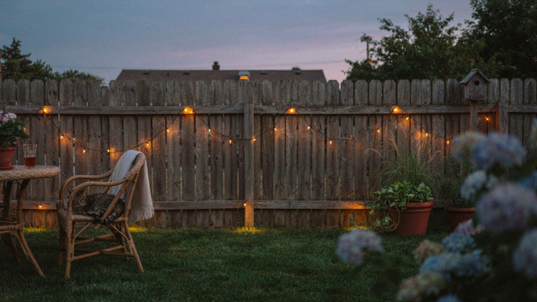 Fairy lights along a fence glow softly in a charming backyard at dusk.