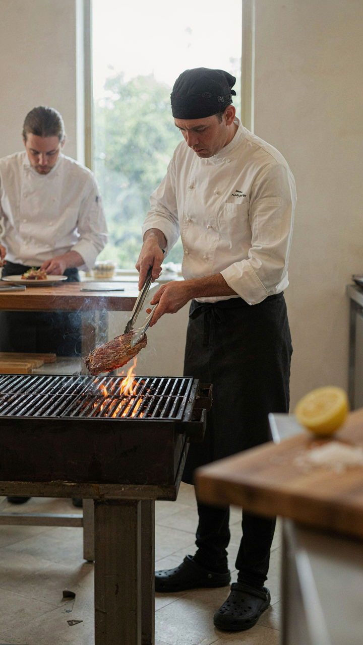 A chef turns food directly over an open flame on a grill.
