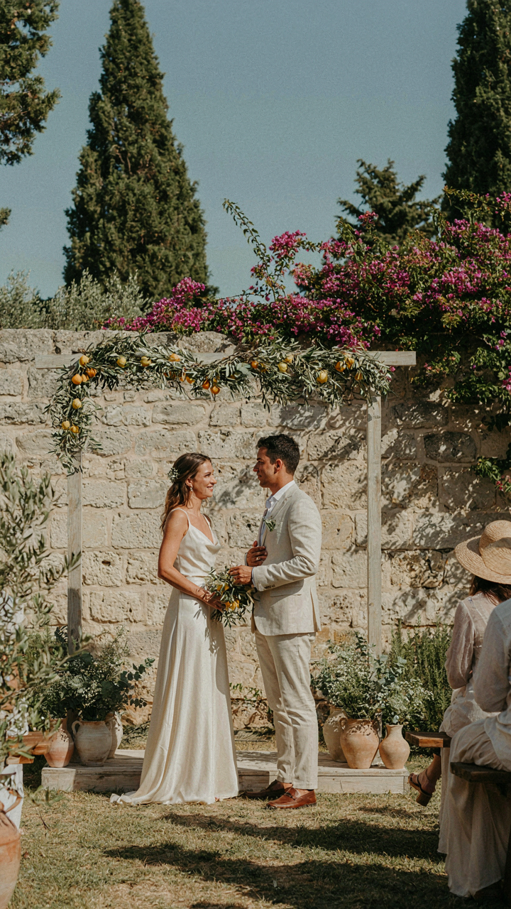 A couple exchanges vows under an arch with olive branches in a Mediterranean garden.