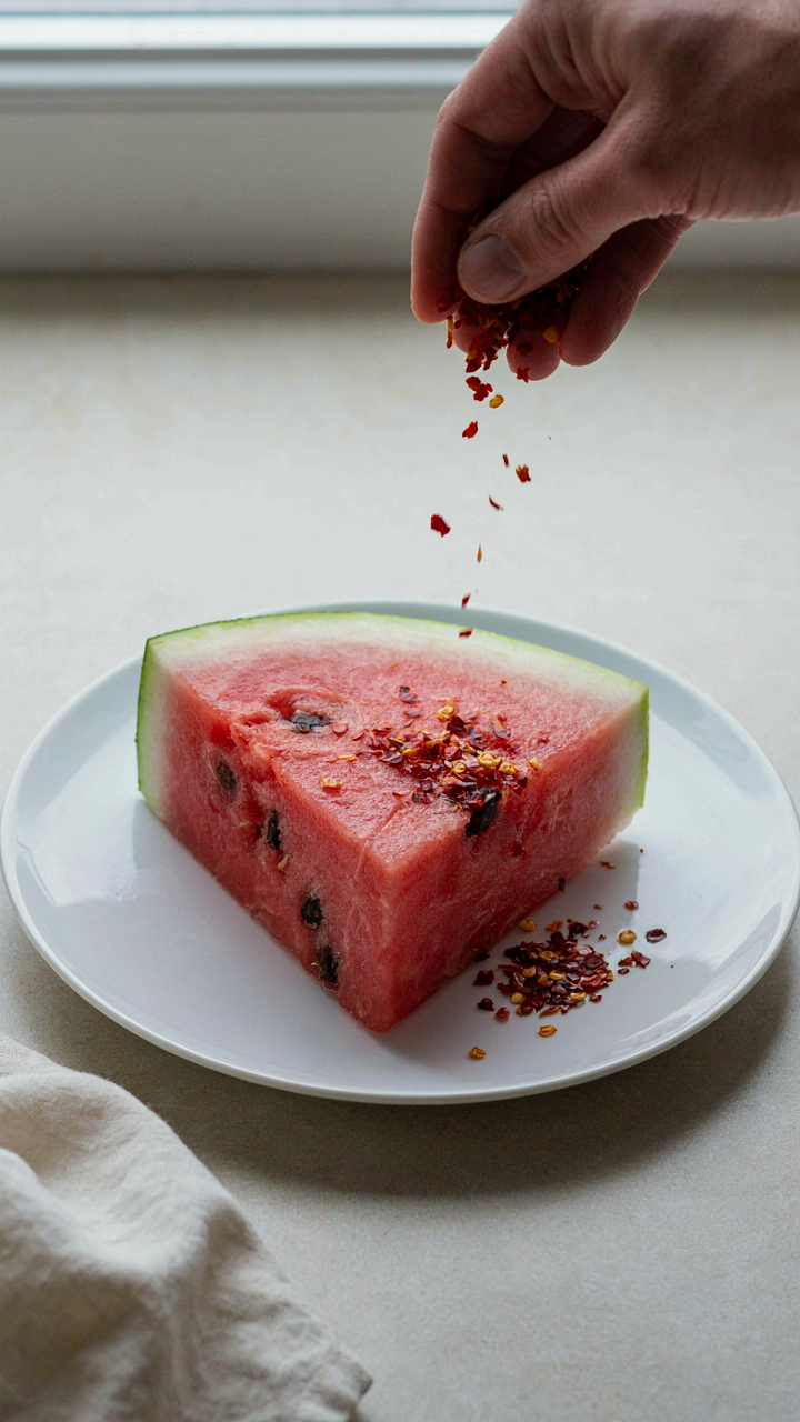 A hand sprinkles red chili flakes over a slice of fresh watermelon on a white plate.