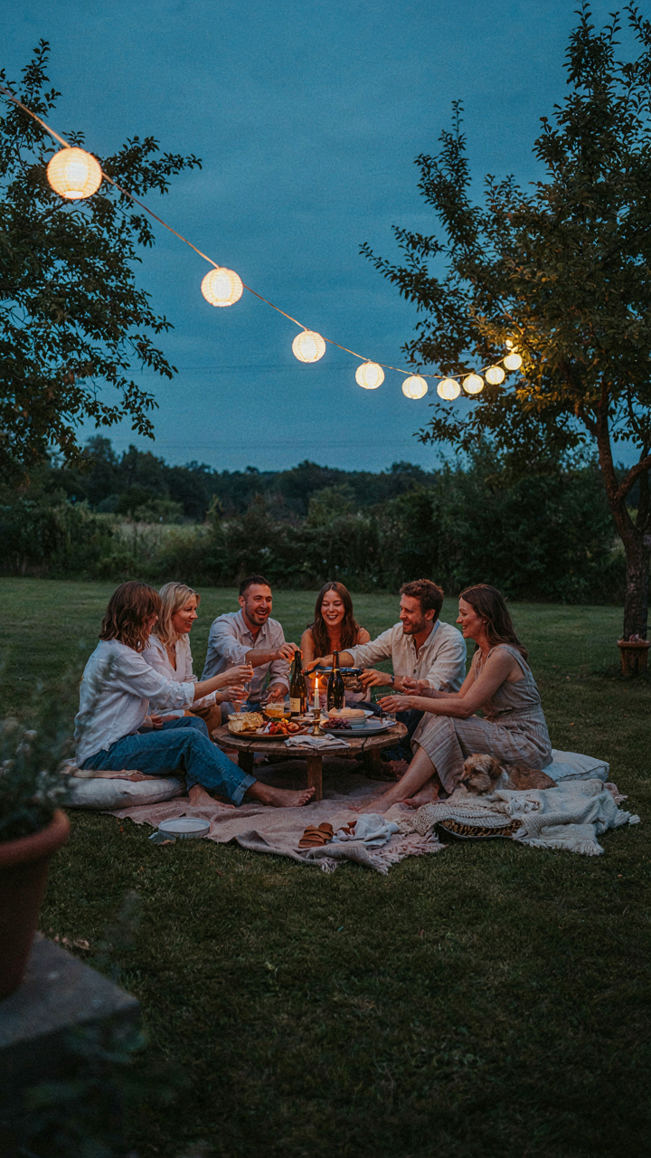 People sit informally on cushions and blankets on a lawn, sharing food under evening lanterns.