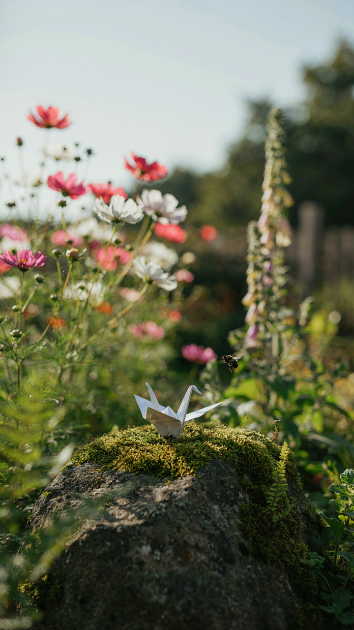 A delicate paper crane rests on a mossy rock in a vibrant garden.