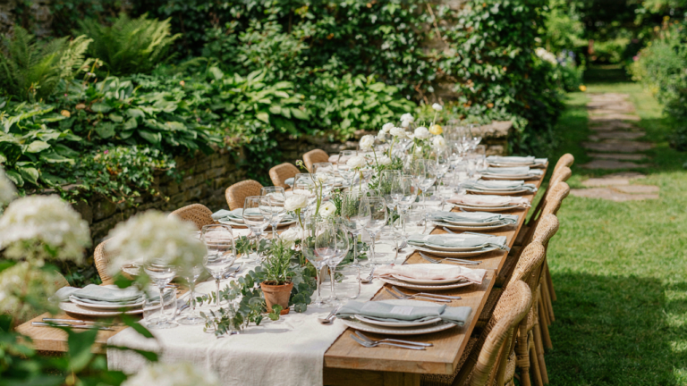A long outdoor table for 12 set with plates and glasses in a garden.