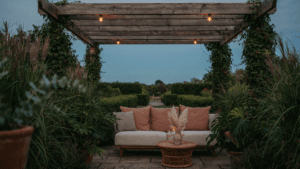 A luxurious pergola seating area glows softly at dusk, surrounded by green plants.