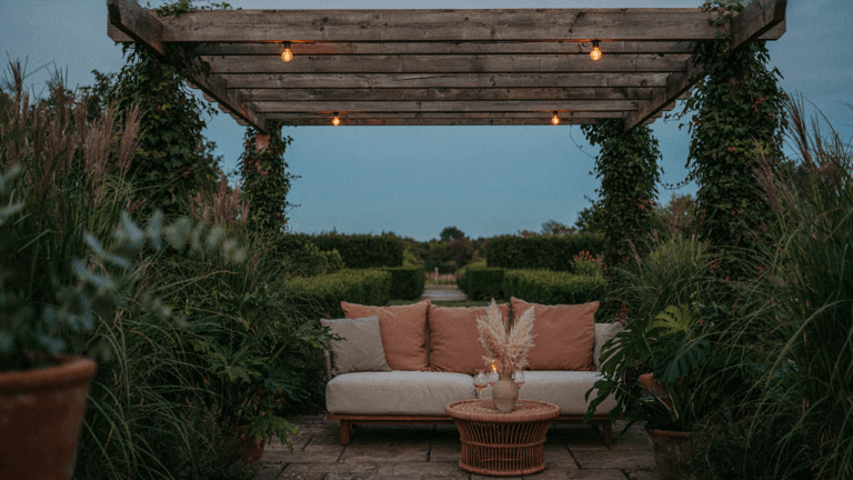 A luxurious pergola seating area glows softly at dusk, surrounded by green plants.