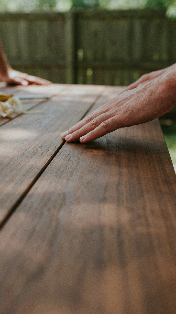 A hand touching the smooth, stained surface of a picnic table.