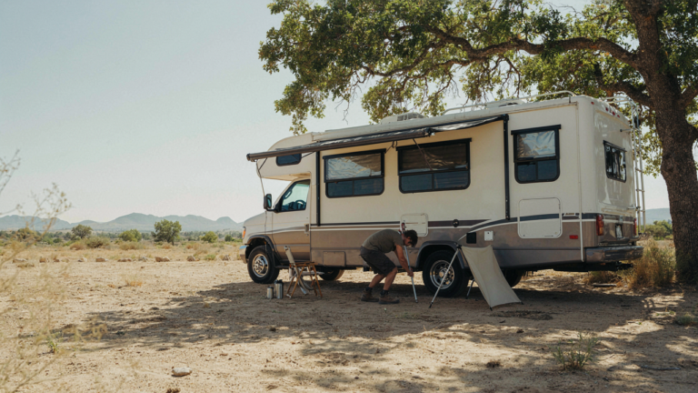 An RV is parked under a large tree with reflective window covers on a sunny day.