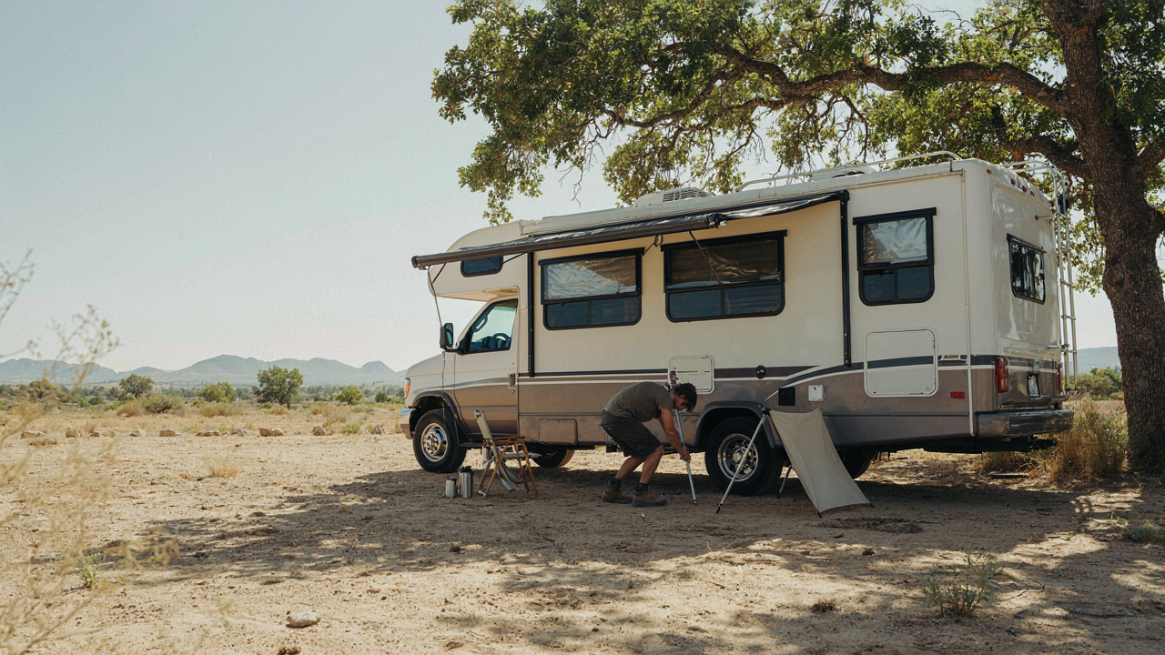 An RV is parked under a large tree with reflective window covers on a sunny day.