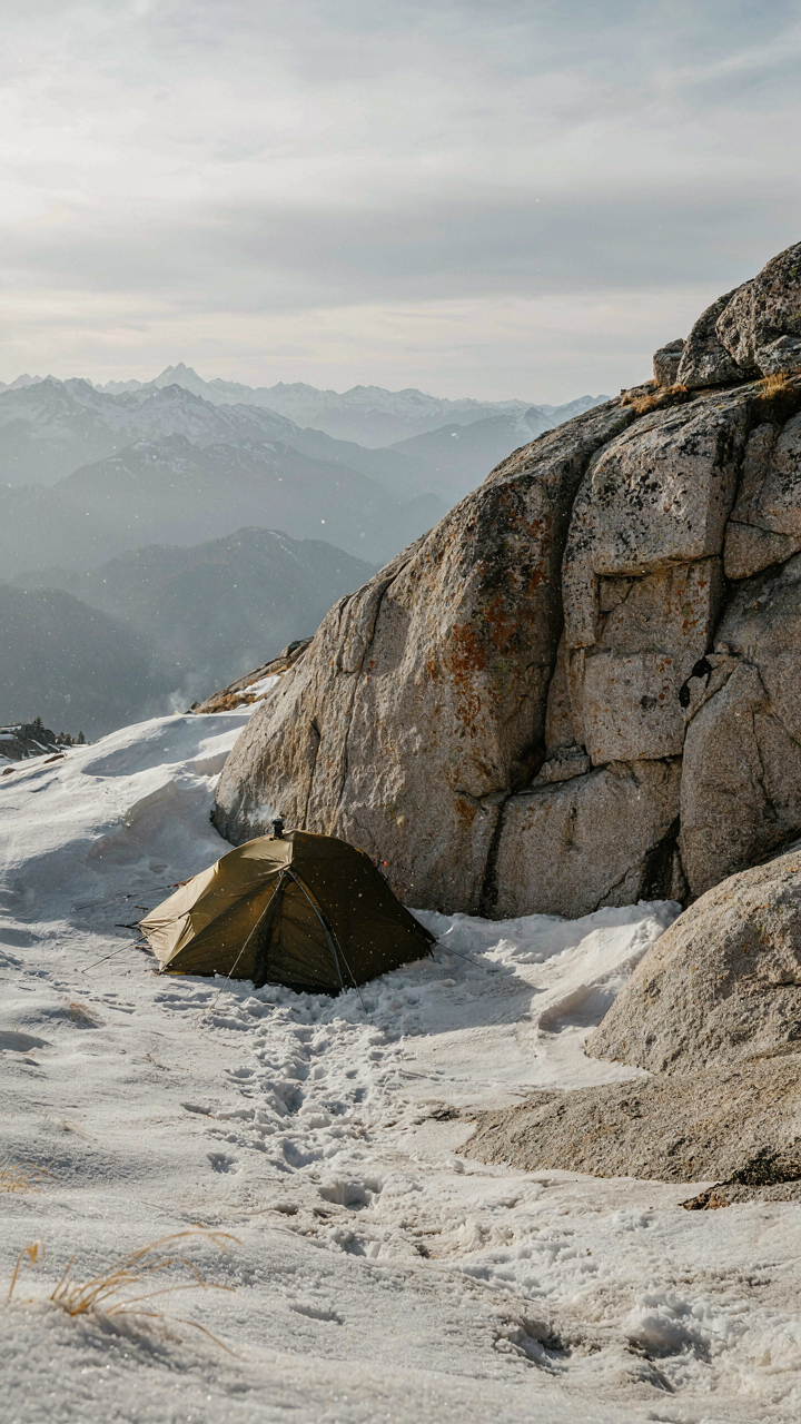 A tent is set behind a big rock on a windy mountain.