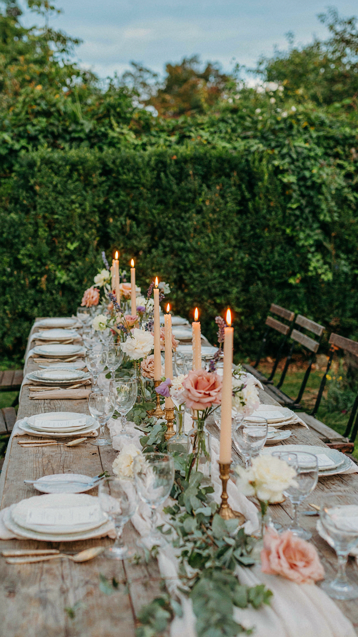 A rustic wedding table with candle centerpieces near a dense hedge.