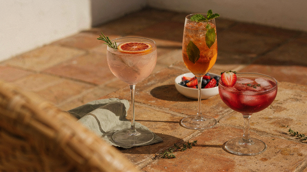Colorful summer spritz mocktails on a table with fresh fruit and herbs in bright sunlight.