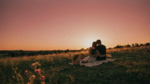 A couple enjoys a sunset picnic on a hillside with a vibrant, colorful sky behind them.