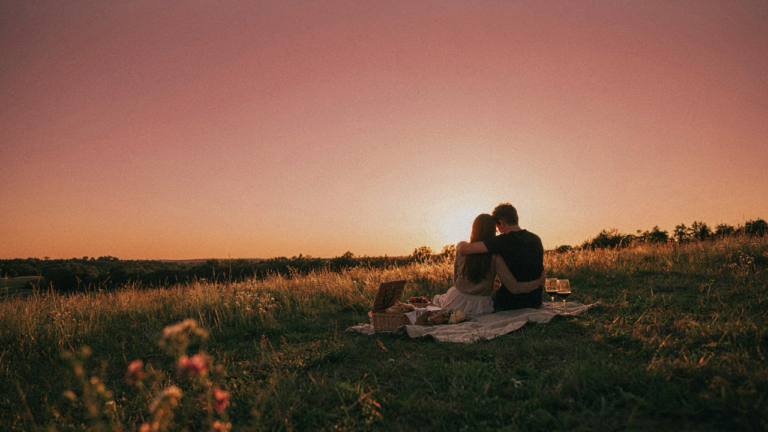 A couple enjoys a sunset picnic on a hillside with a vibrant, colorful sky behind them.