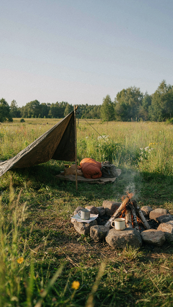 A minimalist camping setup with a tarp, sleeping bag, and campfire in a meadow.