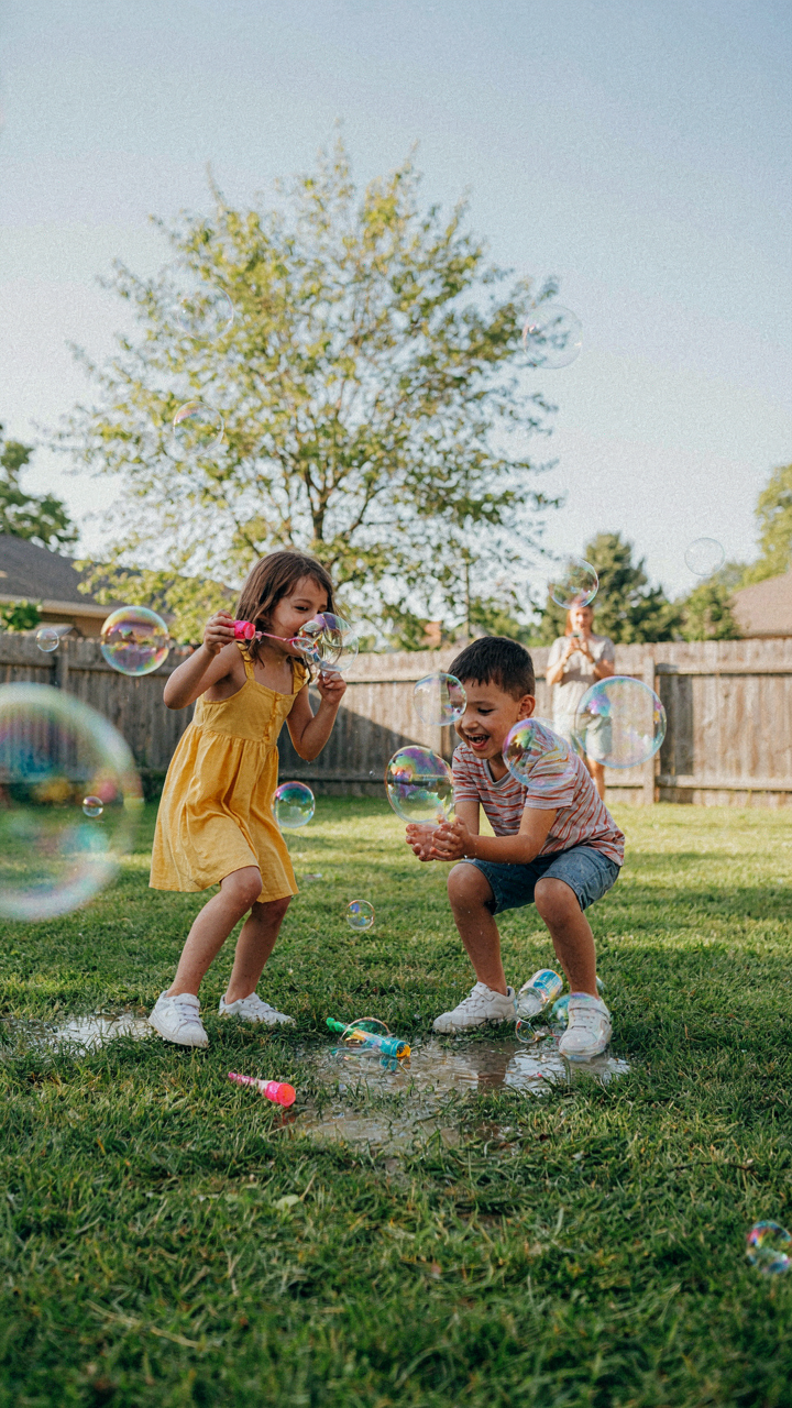 Children giggle and play with oversized bubbles near puddles at a backyard party.