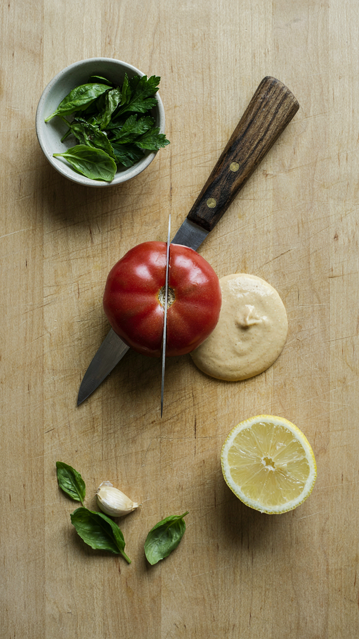 A close-up of a sharp knife slicing a tomato on a cutting board with fresh ingredients.