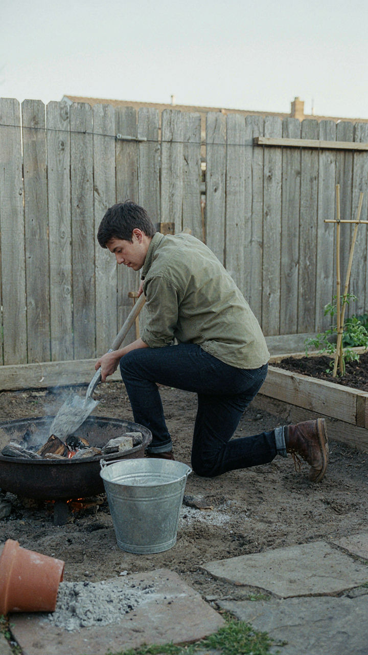 A person shovels wood ash from a fire pit into a metal bucket.