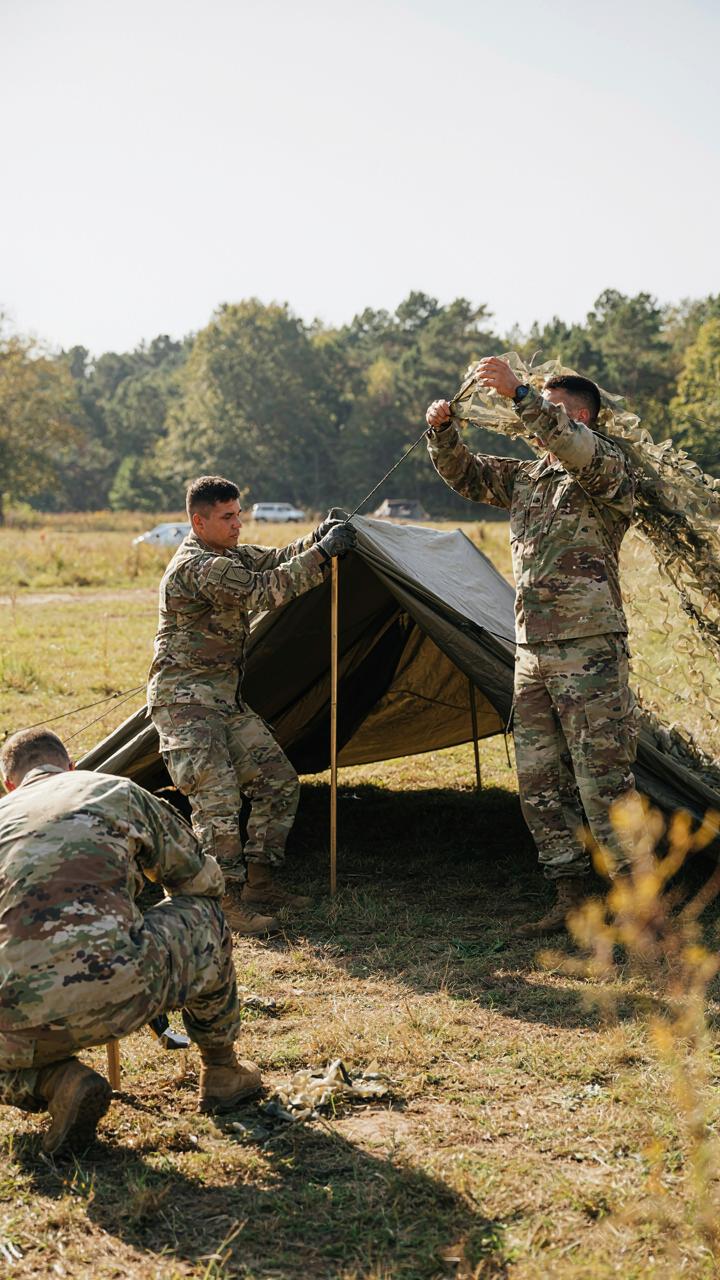 Military personnel position camouflage netting over a tent.