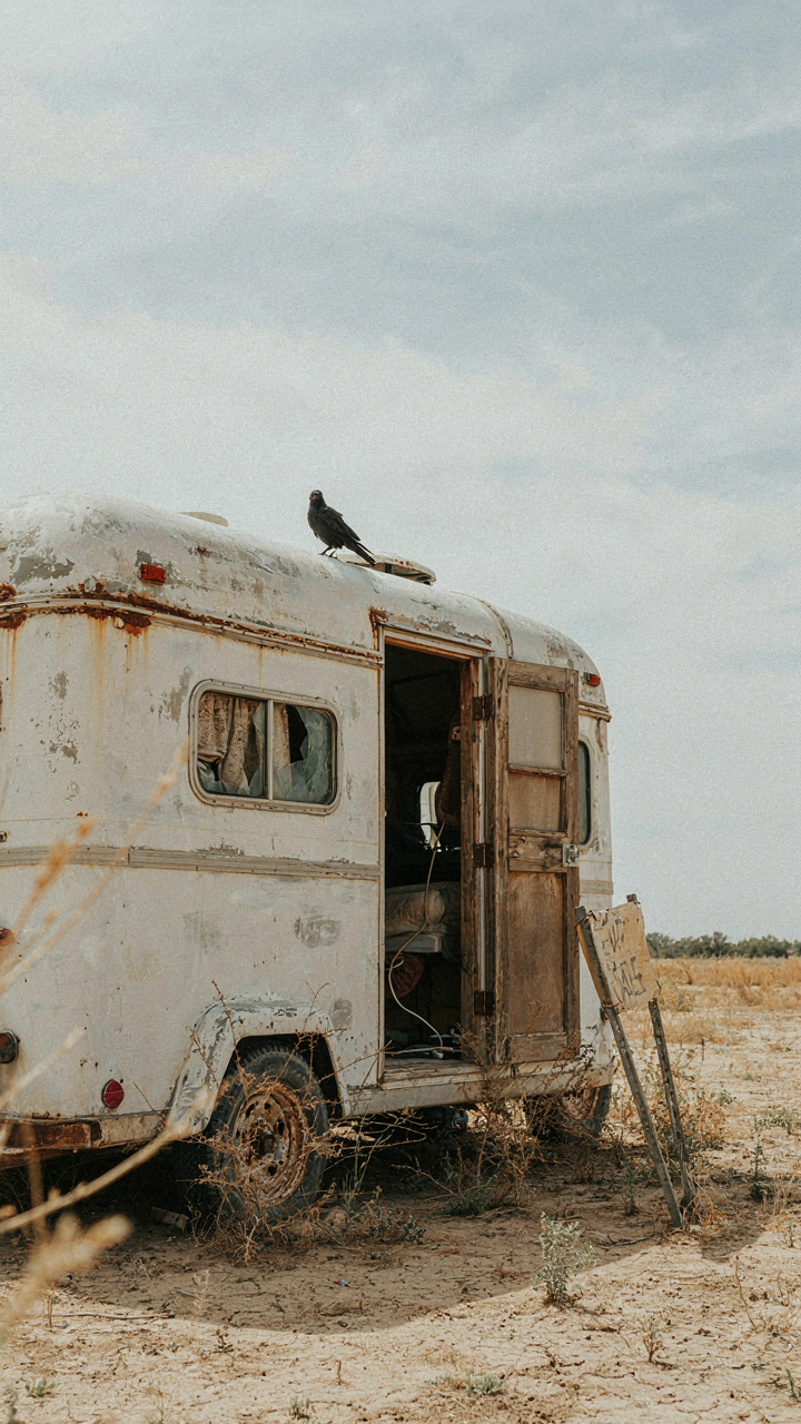 A neglected utility trailer camper conversion in a dusty field.