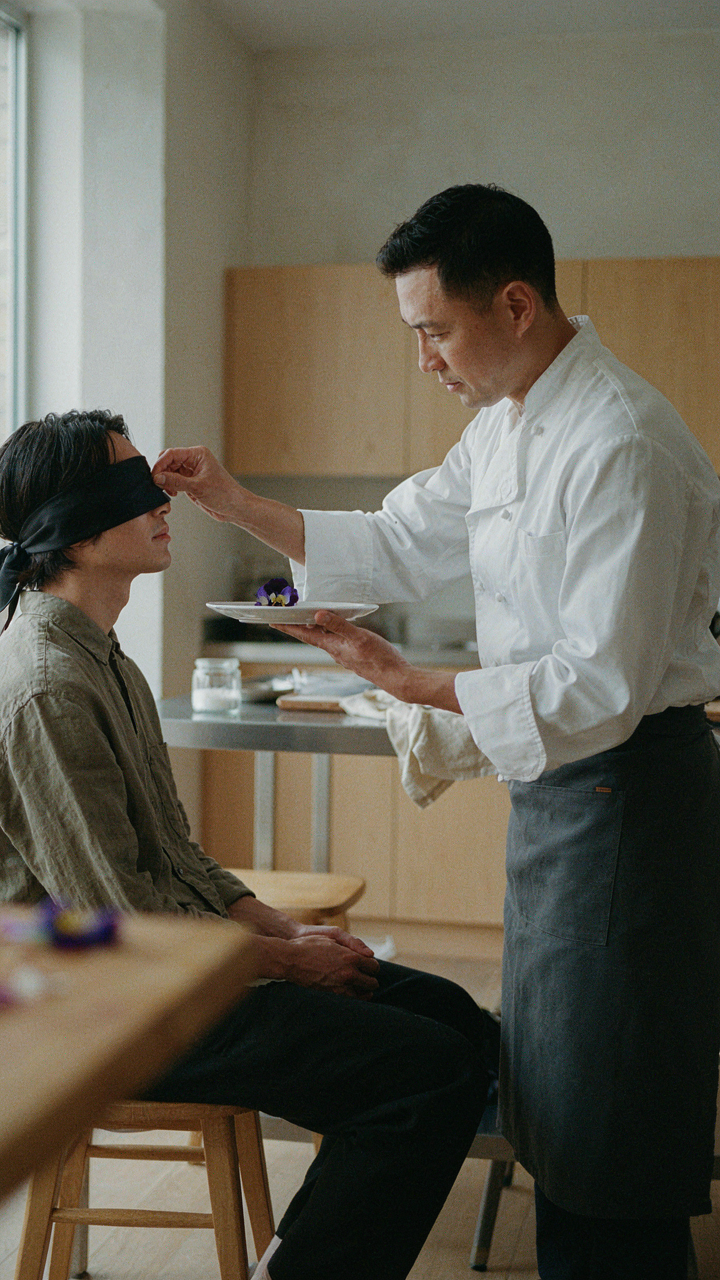 A blindfolded person tastes an edible flower from a small plate, focusing on its flavor.