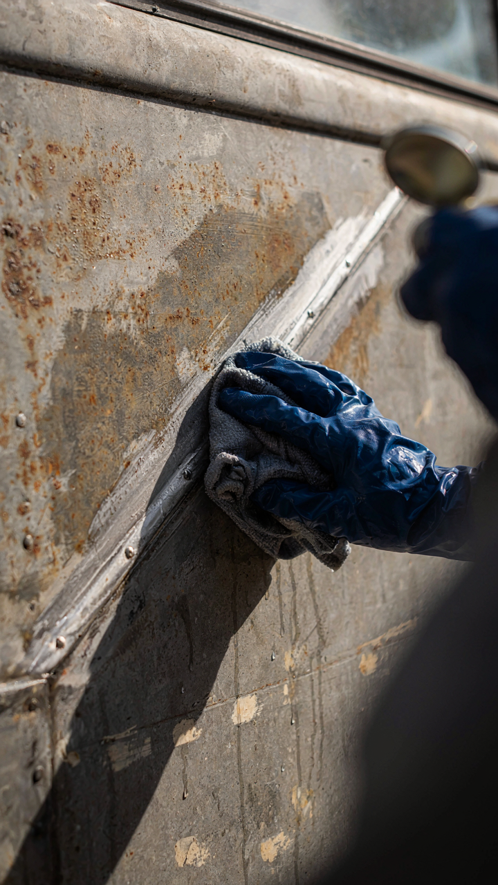 A hand in a glove cleans grease from a vintage camper's metal exterior.