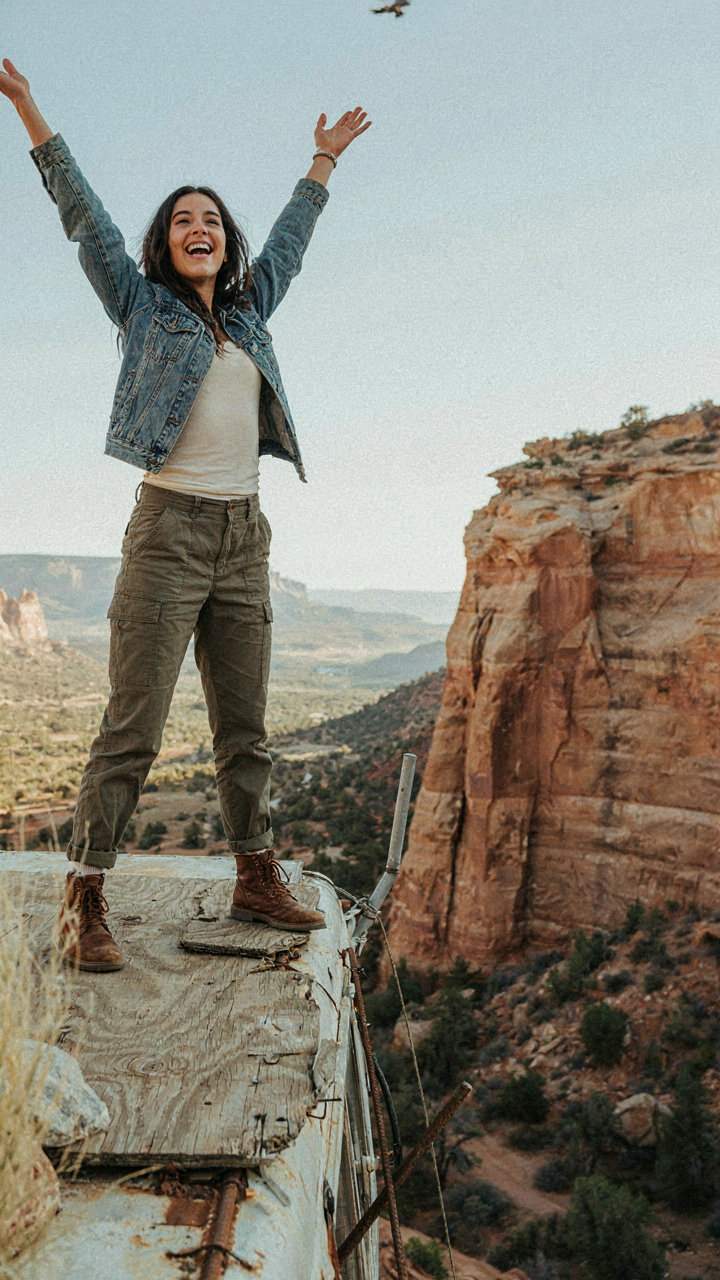 A person stands on a flimsy, unstable skoolie roof deck over a cliff.