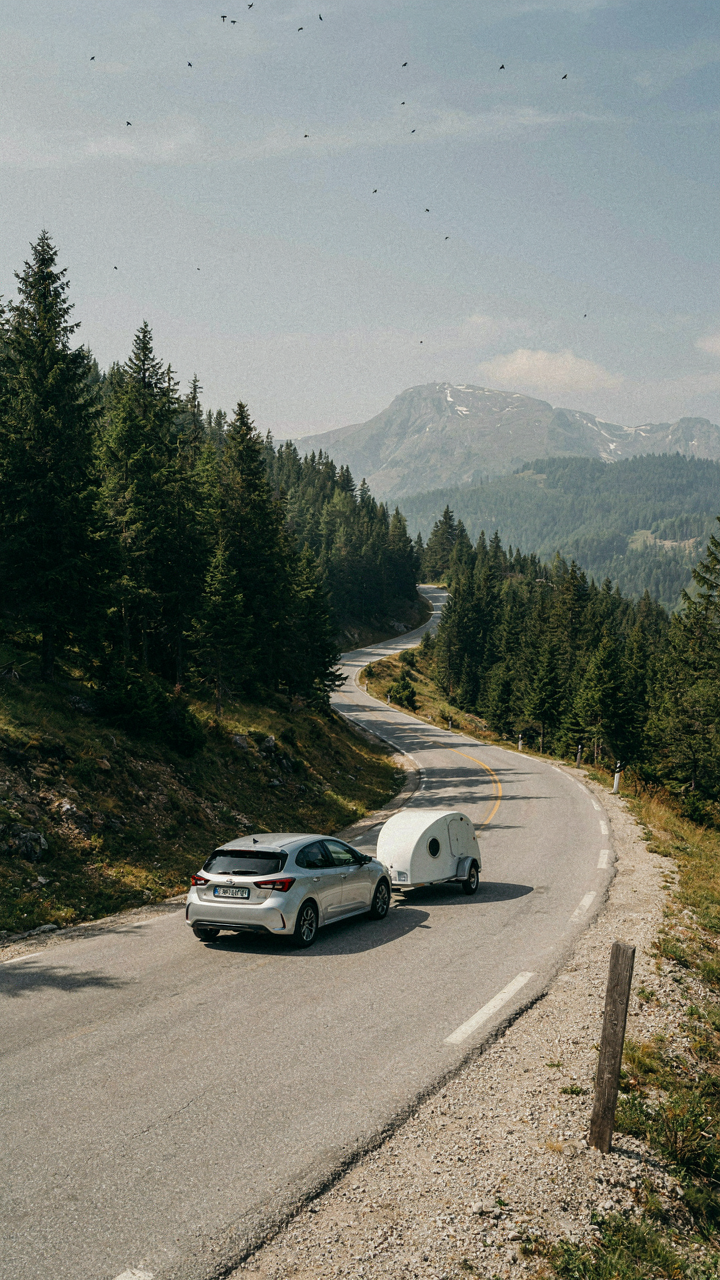 A small camper trailer is pulled by a compact car on a winding mountain road.