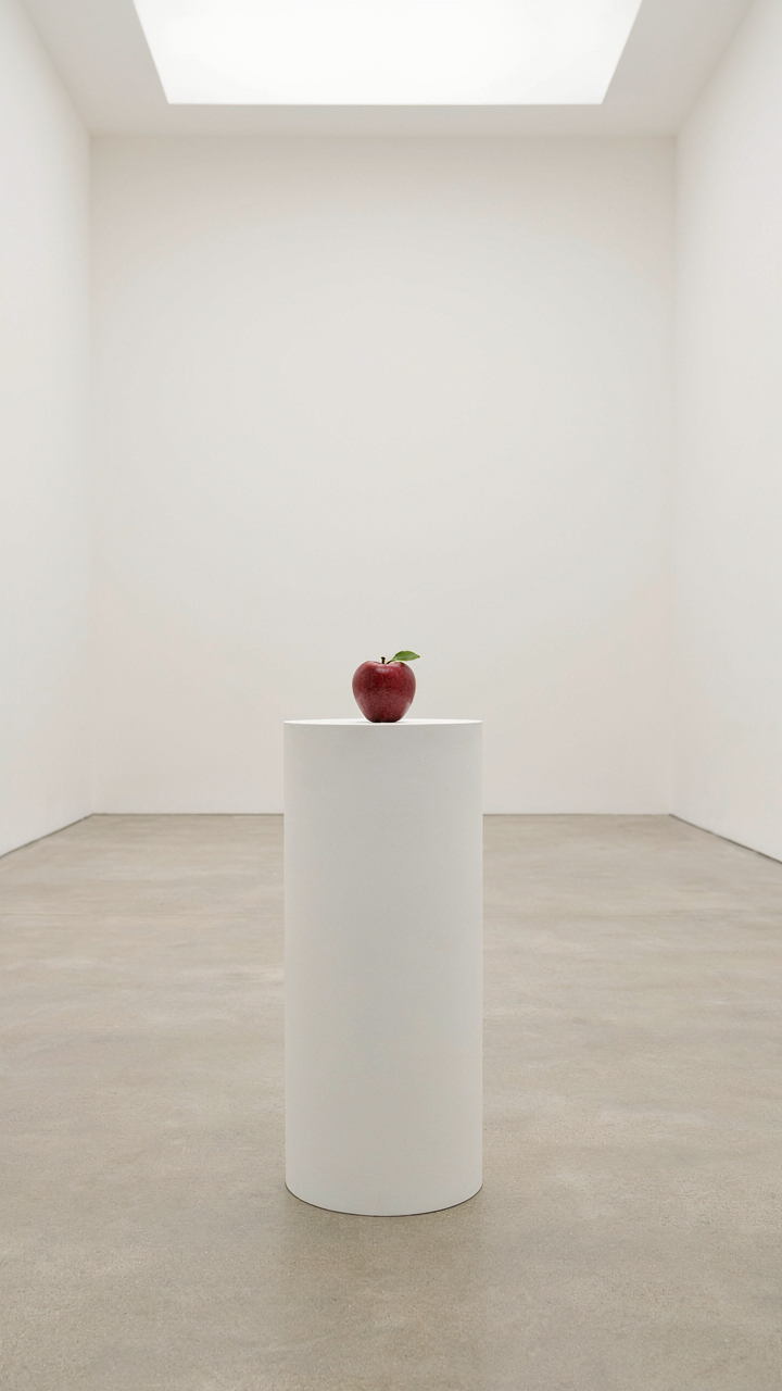 A single red apple on a large white pedestal in an empty art gallery.