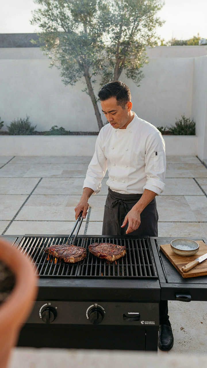 A chef is expertly grilling two steaks on a barbecue in a backyard.