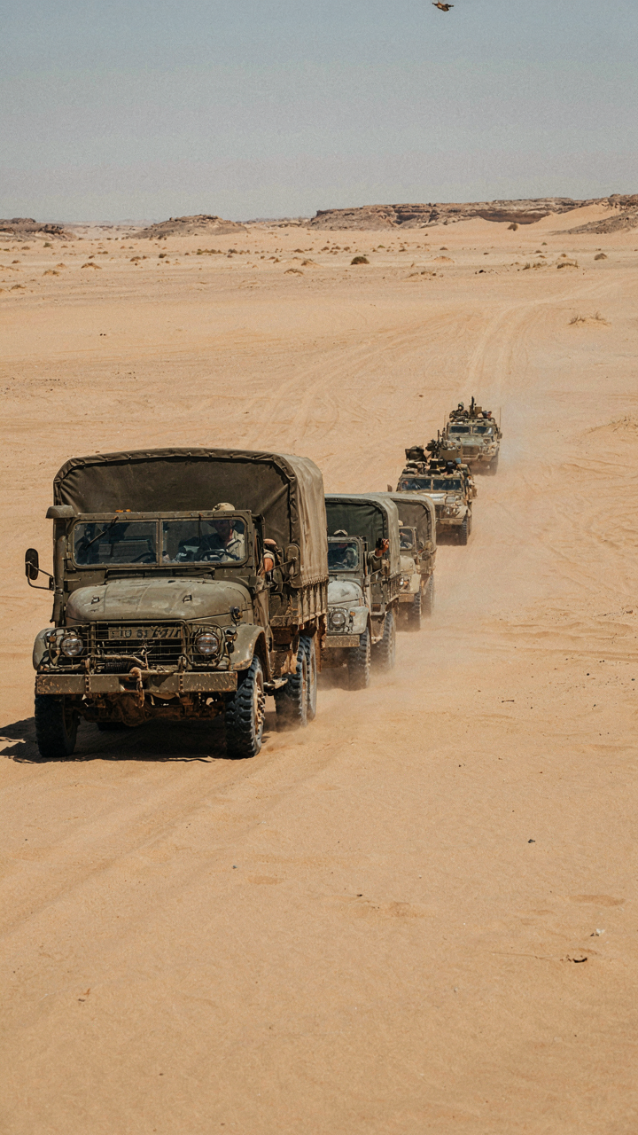 Military vehicles drive in a straight line through the desert.
