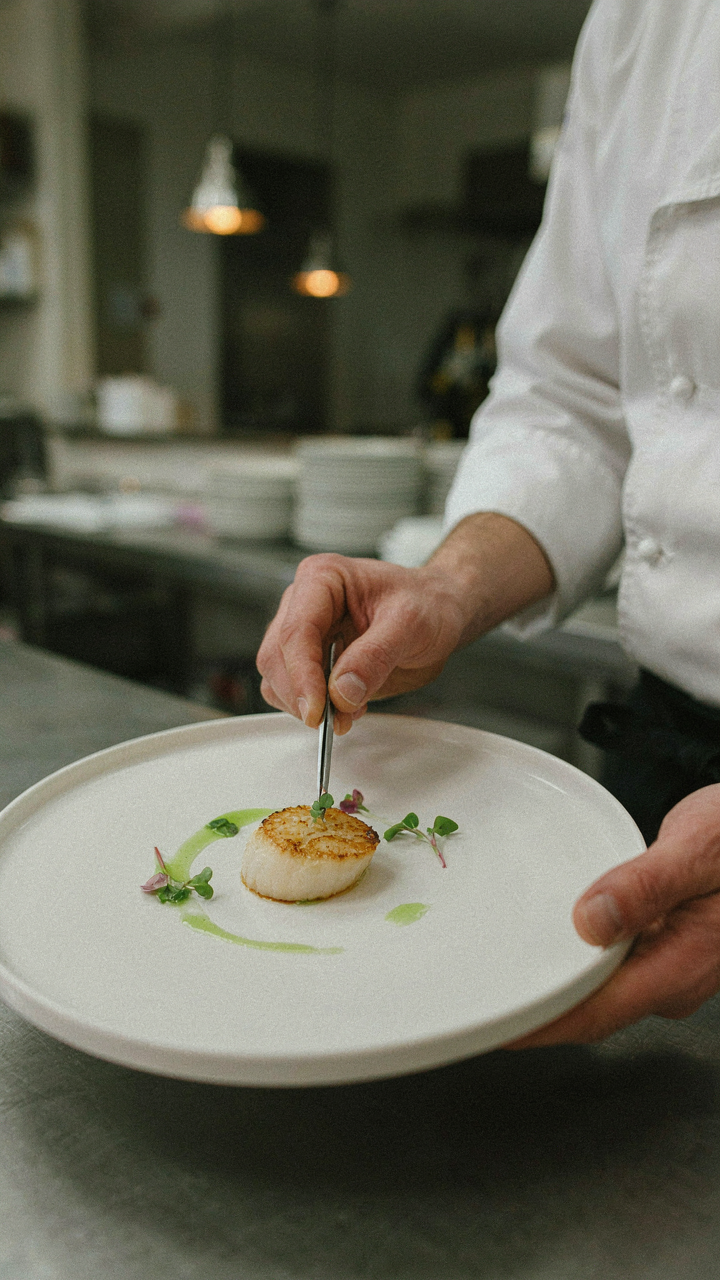 A chef carefully plates a single scallop dish in a professional kitchen.