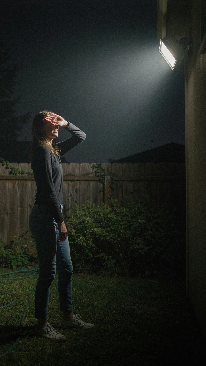A person squints, shielding eyes from a very bright floodlight in a dark backyard.