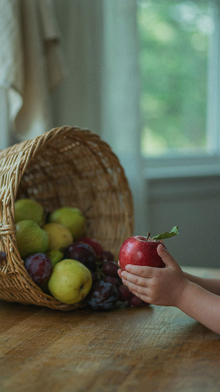 A child holds one red apple, ignoring a full basket of fruit.