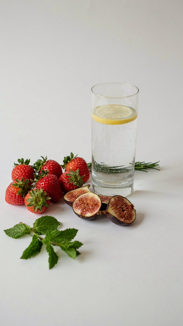 Organic fruits and herbs next to a glass of clear liquid on a white table.