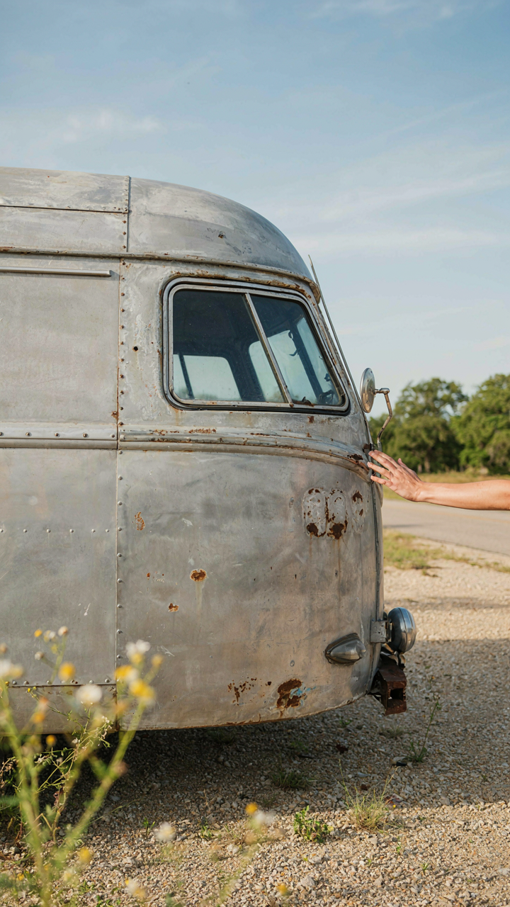 A hand gently touches the weathered metal surface of an old camper.