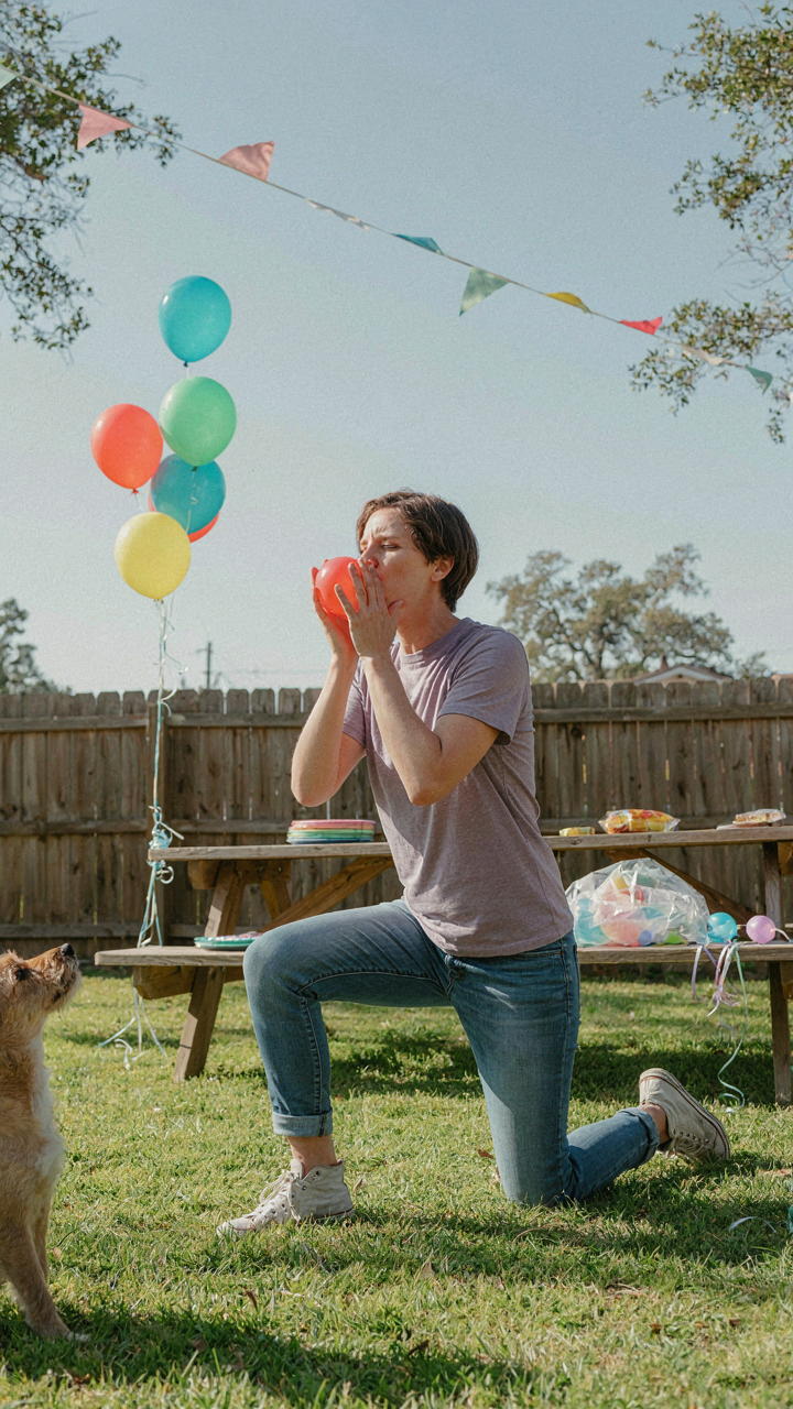 A parent happily blows up balloons by hand in a backyard.
